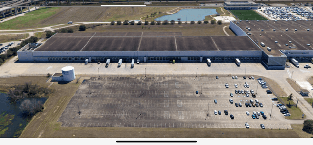 Aerial view of a large warehouse with loading docks and a vacant parking lot. Nearby, a pond and additional industrial buildings are visible.