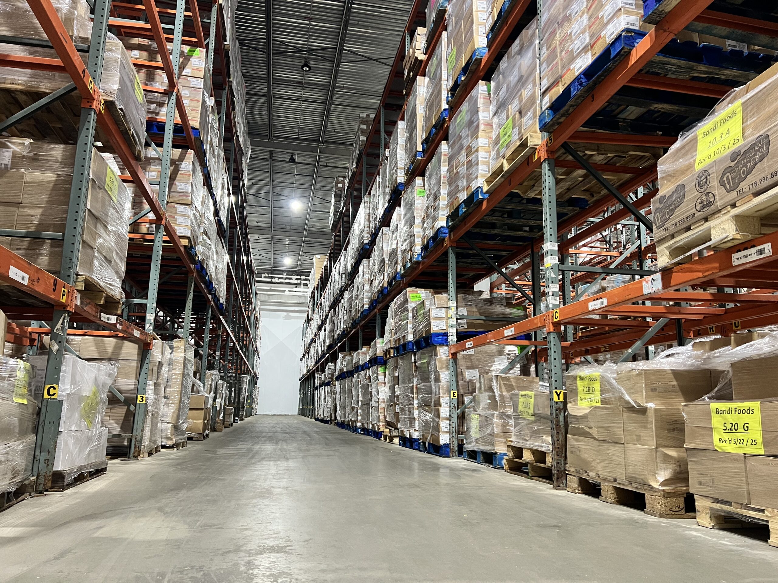 Warehouse aisle lined with tall shelving units stacked with boxes and pallets. Brightly labeled products suggest an organization of inventory.