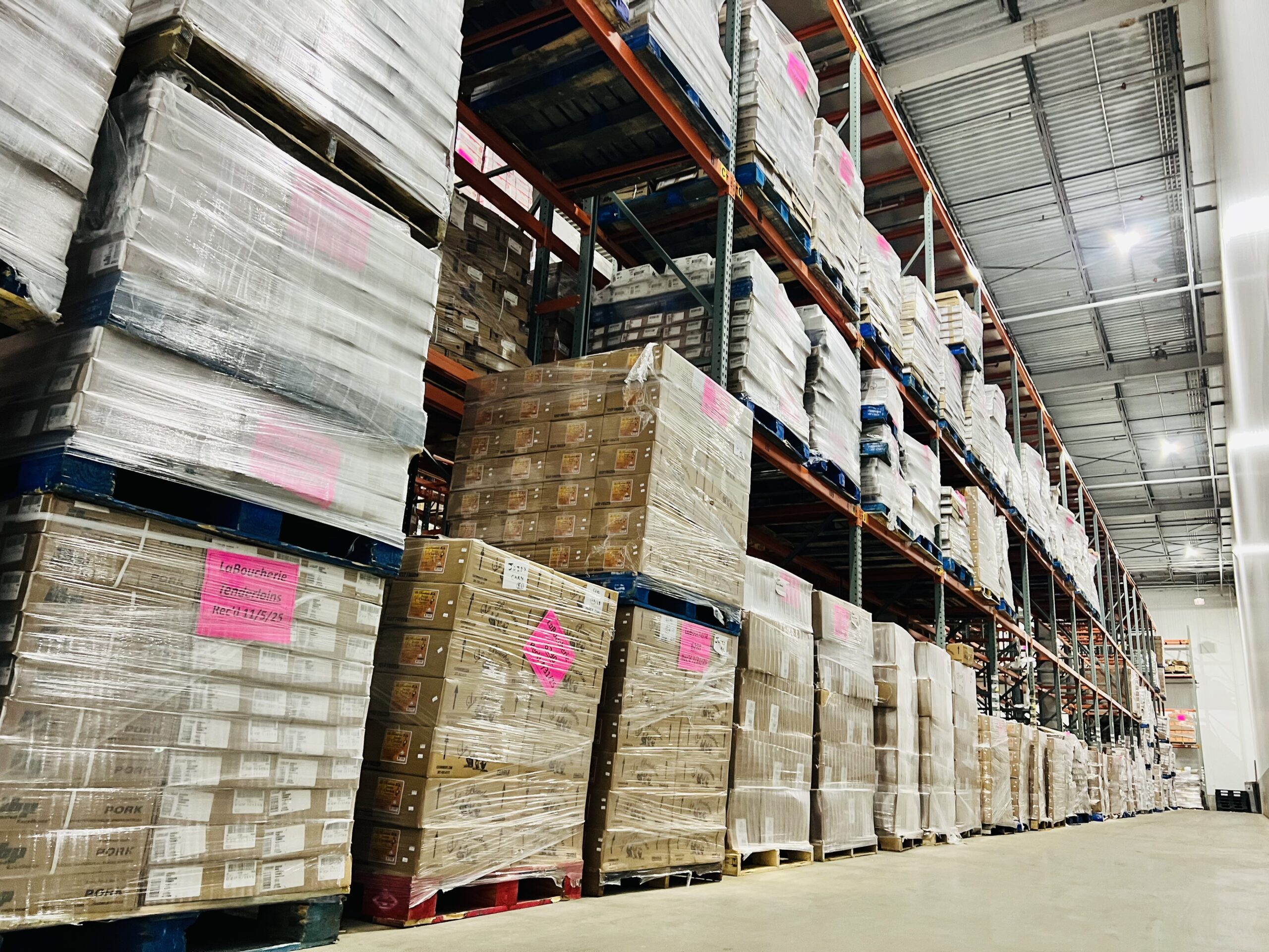 Warehouse interior with stacked pallets of products, labeled with pink tags. The image highlights inventory organization and storage efficiency.