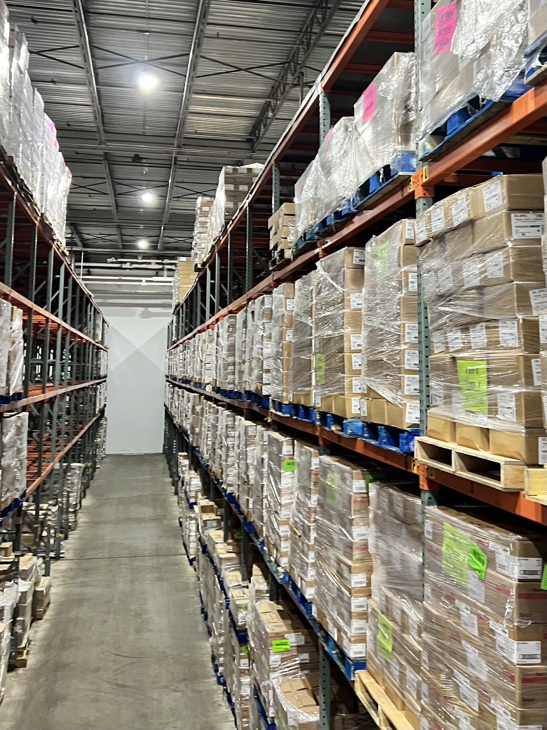 Warehouse aisle lined with tall shelving units stacked with palletized goods. Bright lights illuminate the organized space, highlighting inventory management.