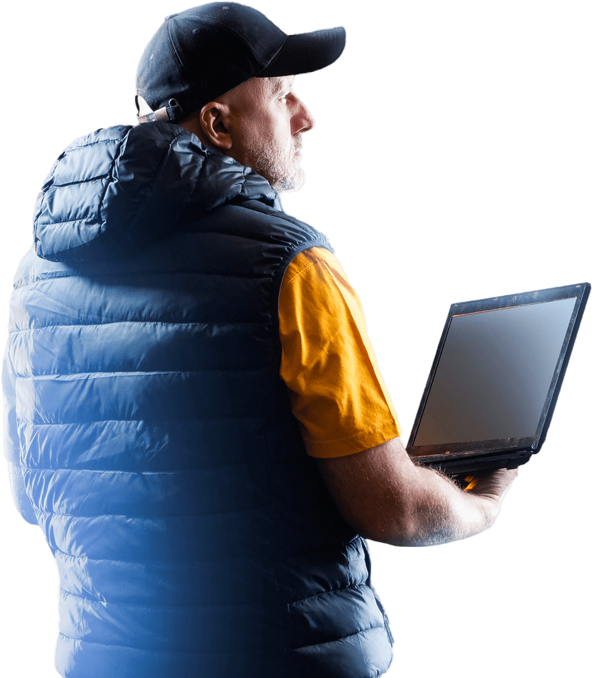 Man in a black puffer vest and cap holds an open laptop, looking thoughtfully to the side, suggesting a tech or work-related theme.
